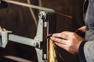 a person holding a piece of metal up to a belt grinder with sparks coming off